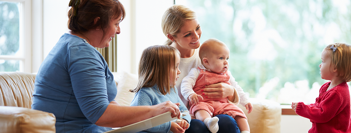 A health visitor with a clipboard consults with a mother and her three young children in a bright, comfortable living room.